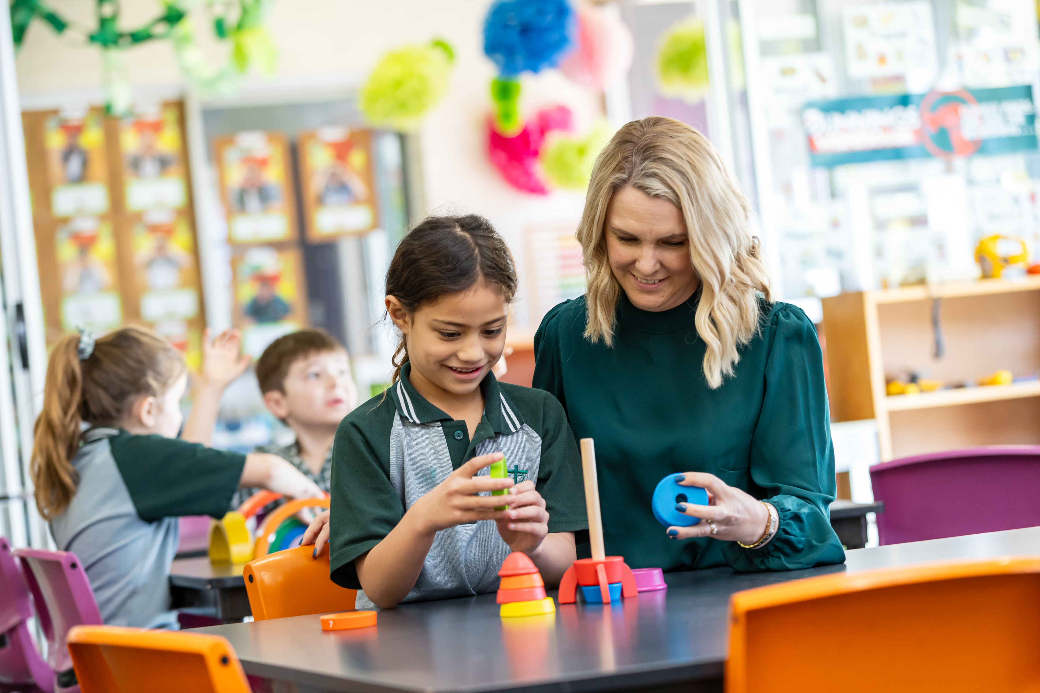 Adult supporting a student using colourful stacking toys at a classroom table, with other students working in the background.
