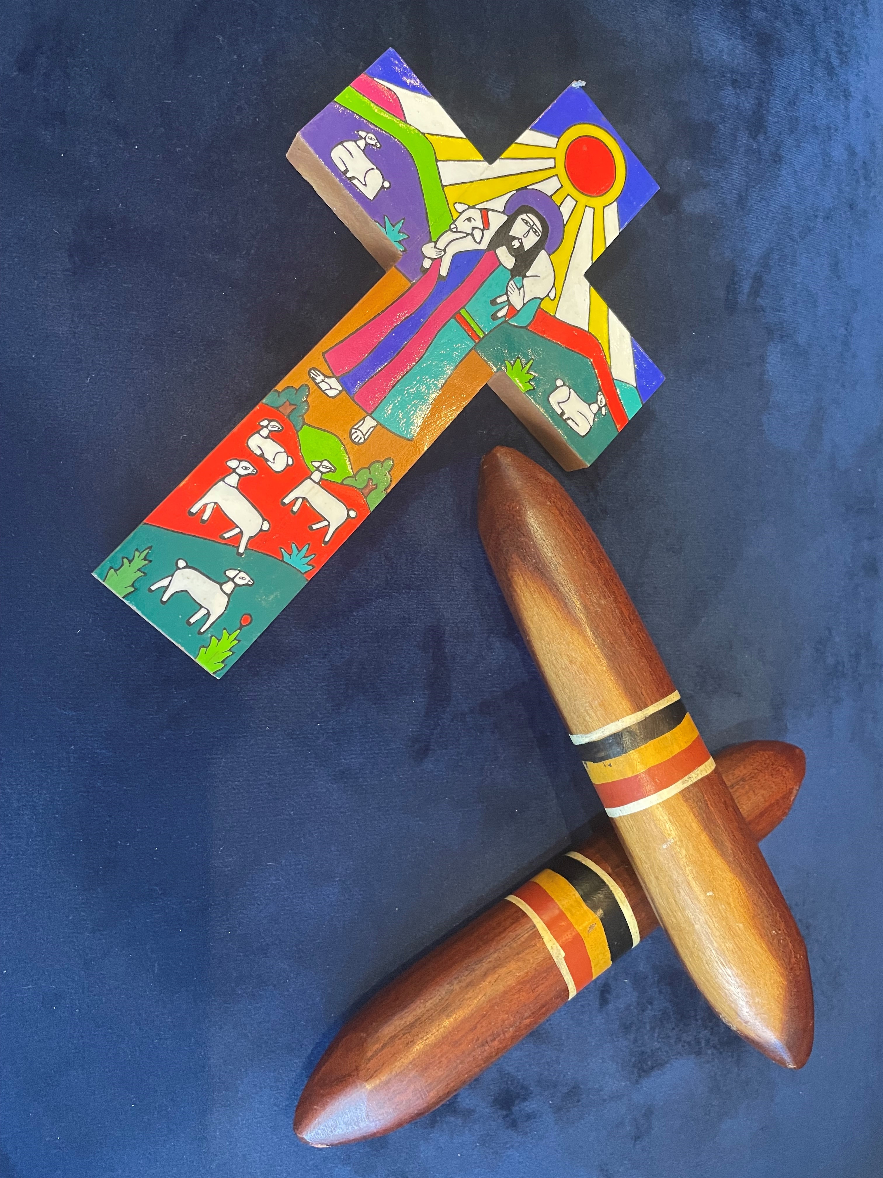 Decorated cross with colourful painted scenes placed beside two wooden clapping sticks on a dark surface.