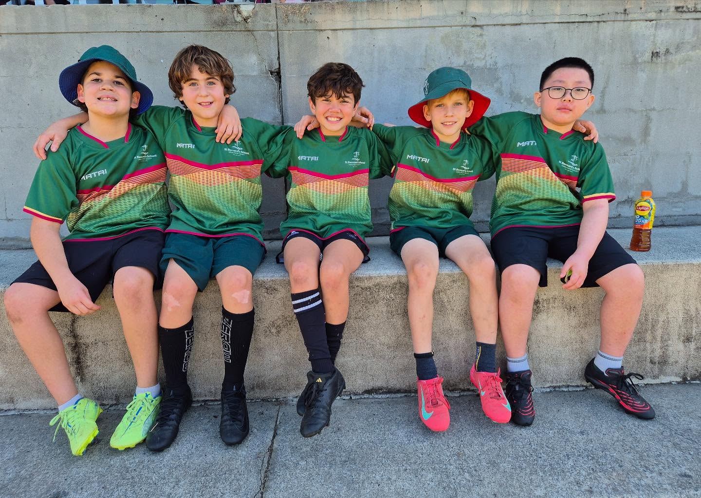 Five students in green sports uniforms sitting together on a bench with arms around each other outdoors.