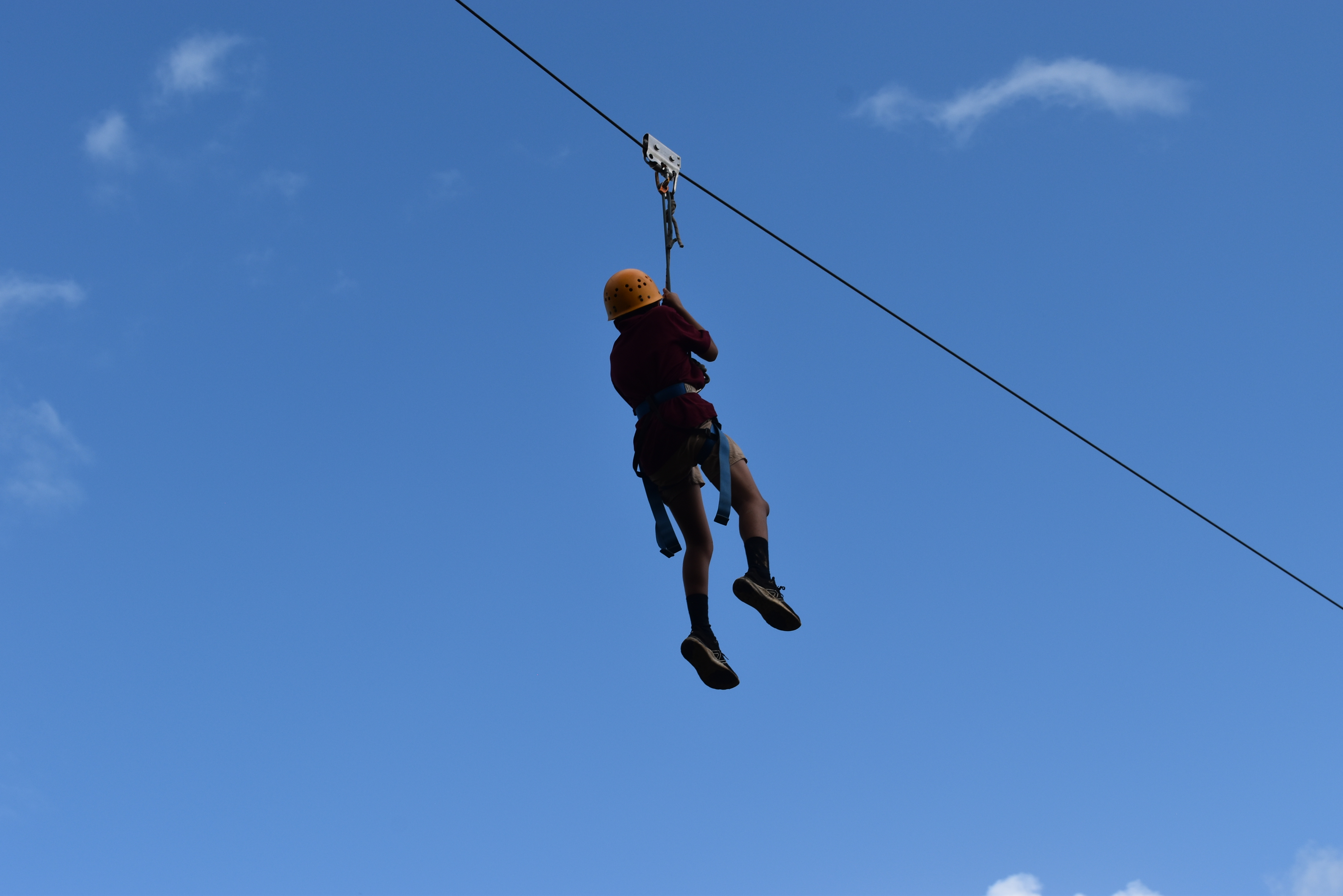 Student wearing a helmet and harness travelling along a zipline against a blue sky.