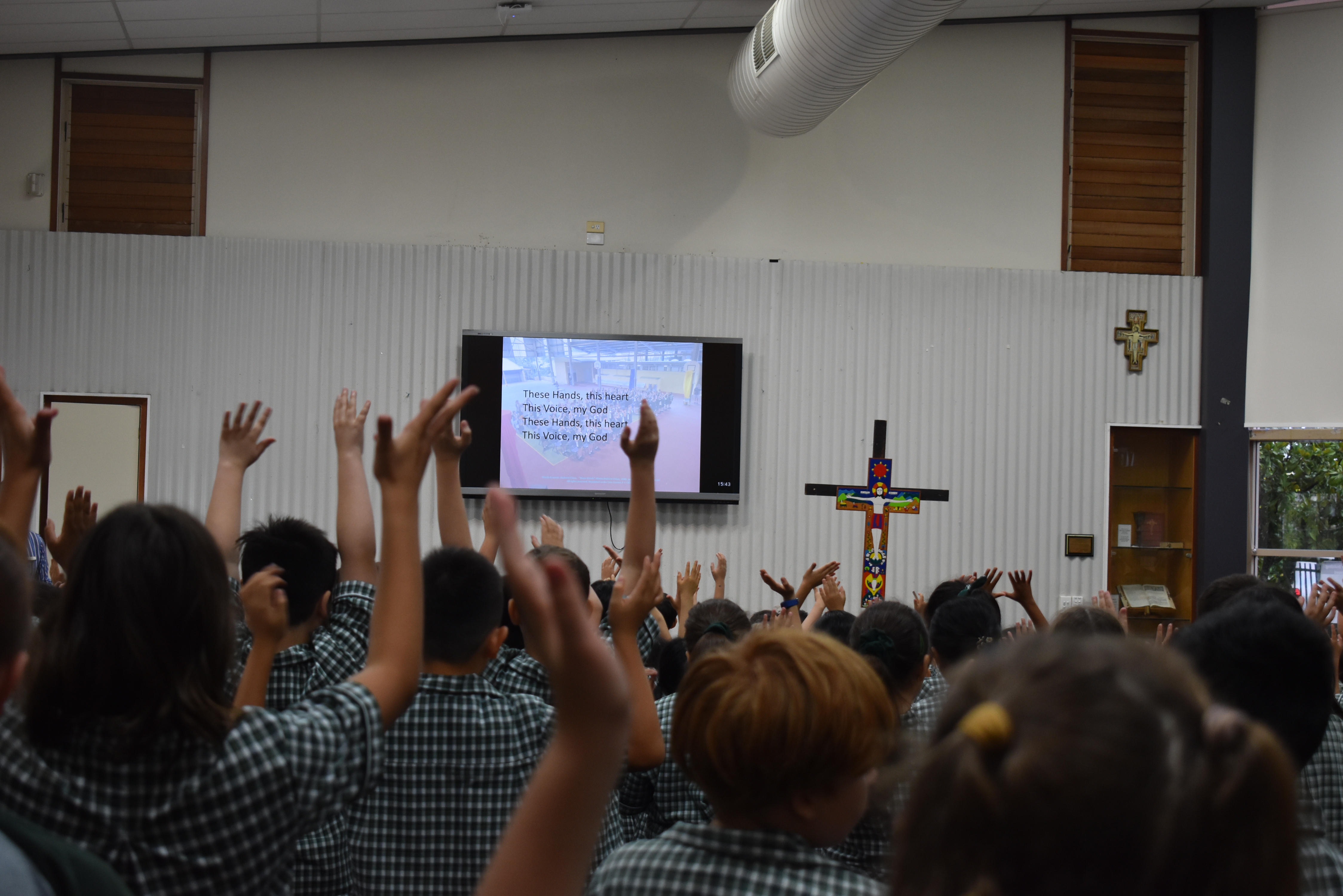 Students gathered in a school hall with hands raised during a liturgy, with a screen displaying song lyrics and a cross on the wall.