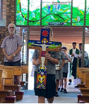 Student in a church holding a decorated cross during a procession, with others following and stained glass windows in the background.