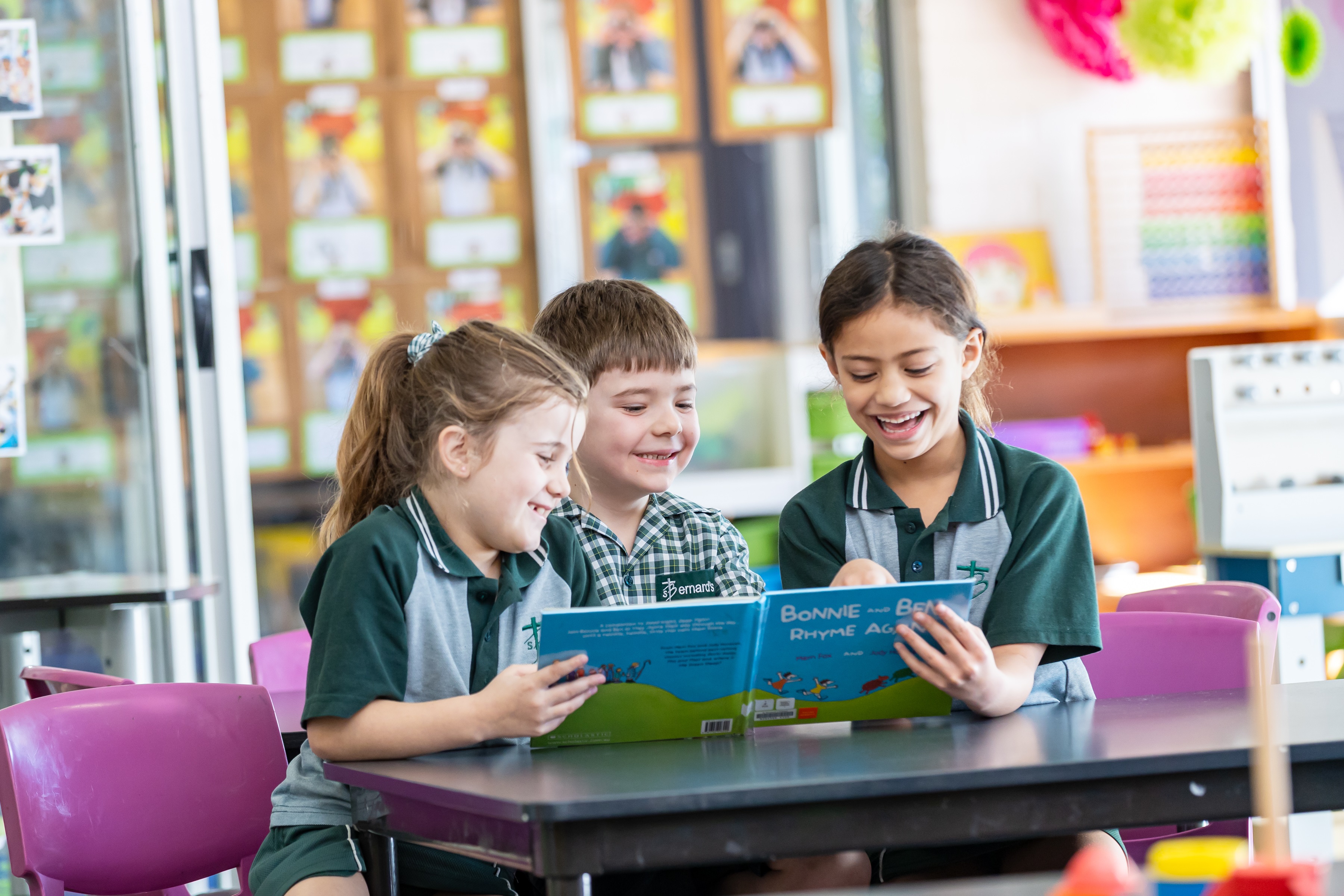 Three students in school uniforms sitting together at a table reading a book in a classroom.