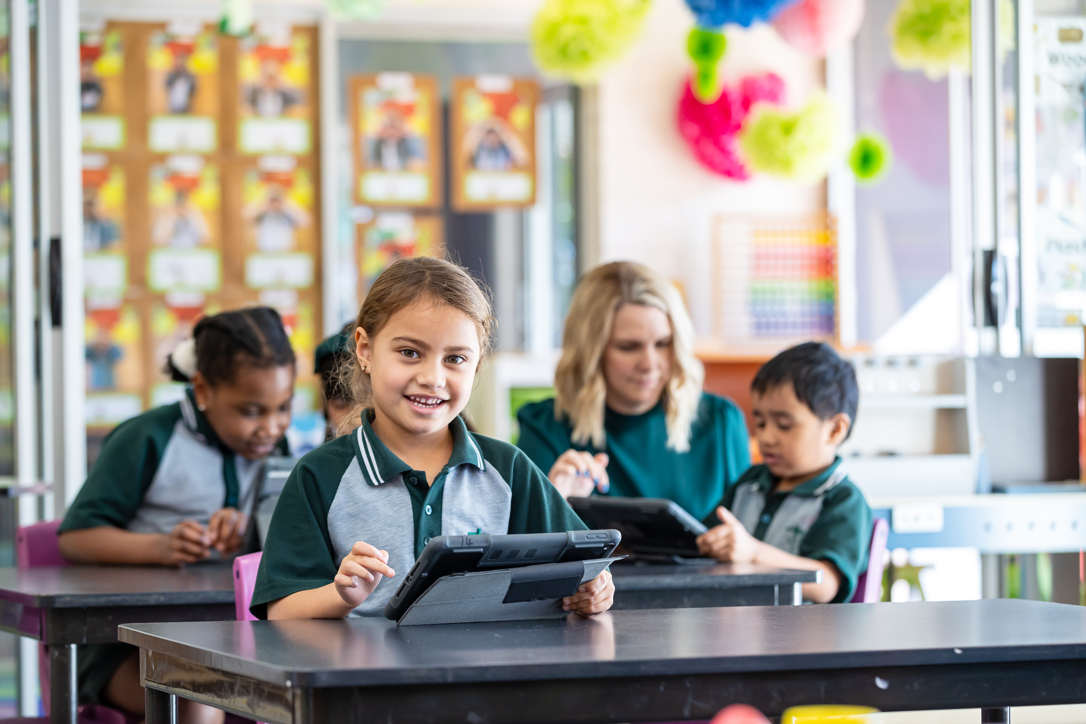 Student using a tablet at a classroom desk, with other students and an adult working on devices in the background.