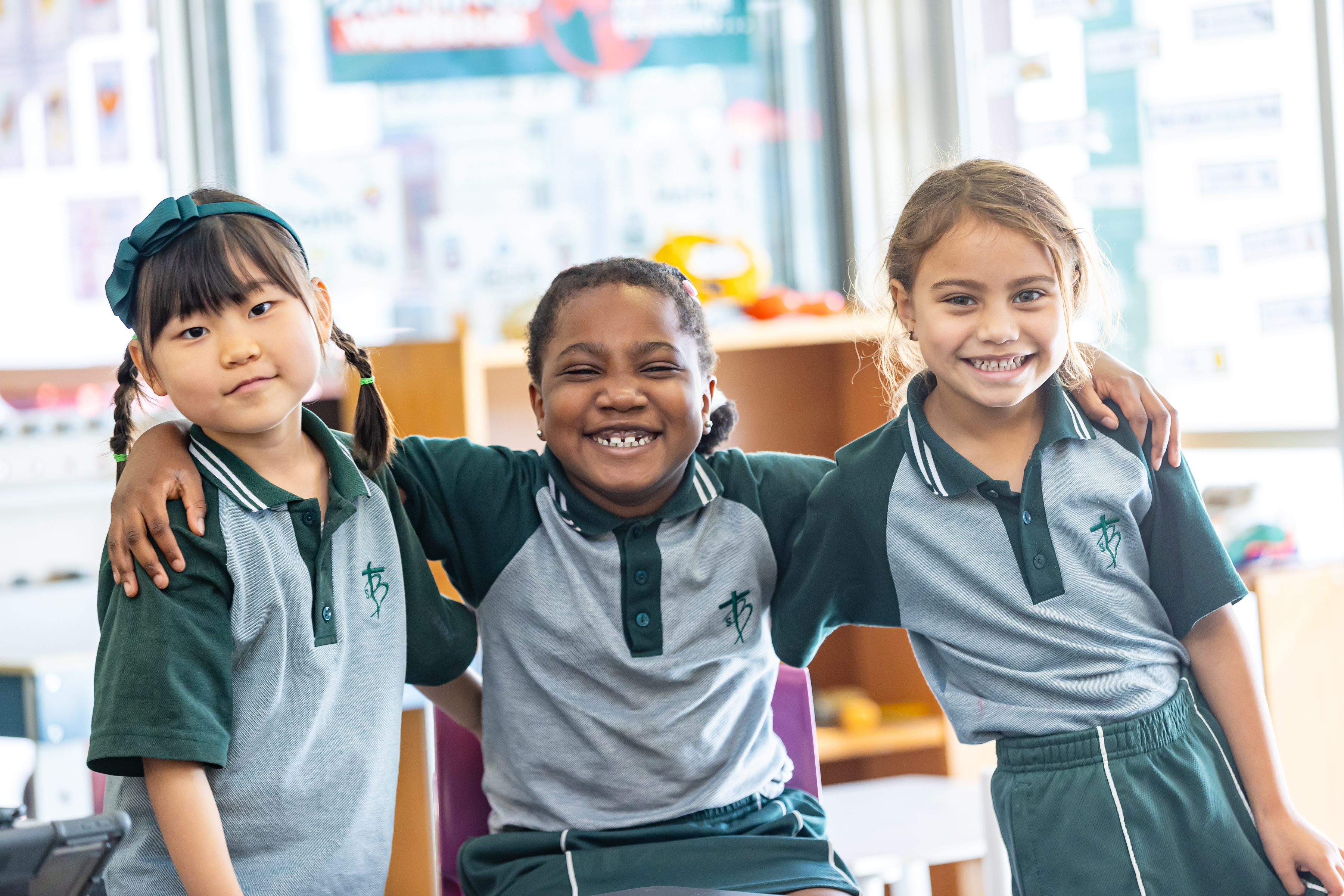Three students in school uniforms standing with arms around each other in a classroom.