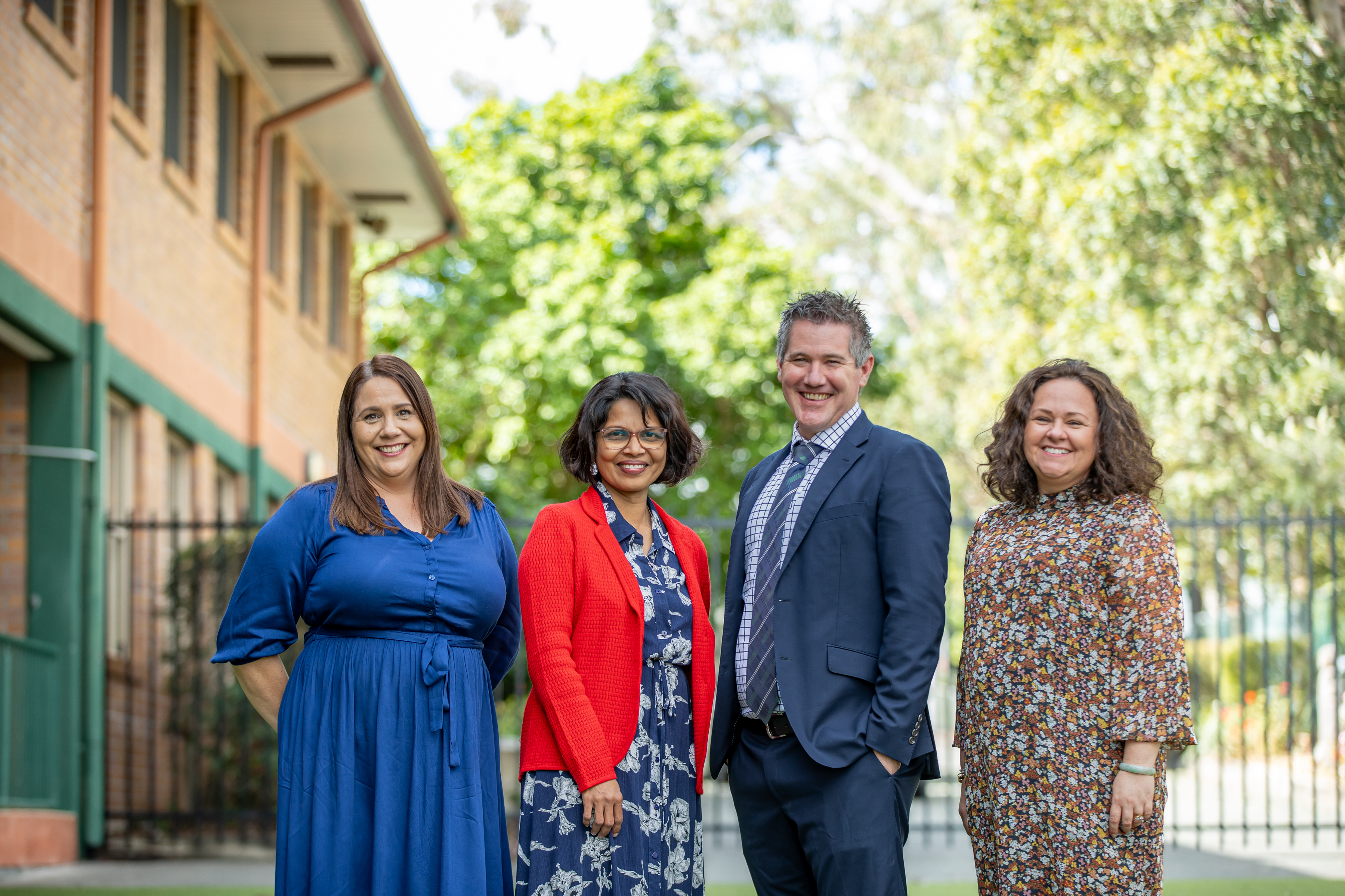 Four adults standing together outdoors on a school campus with a building and trees in the background.