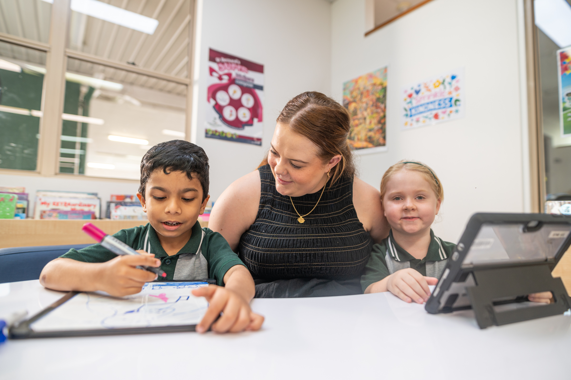 Adult working with two students at a table, guiding a drawing activity while another device sits nearby.