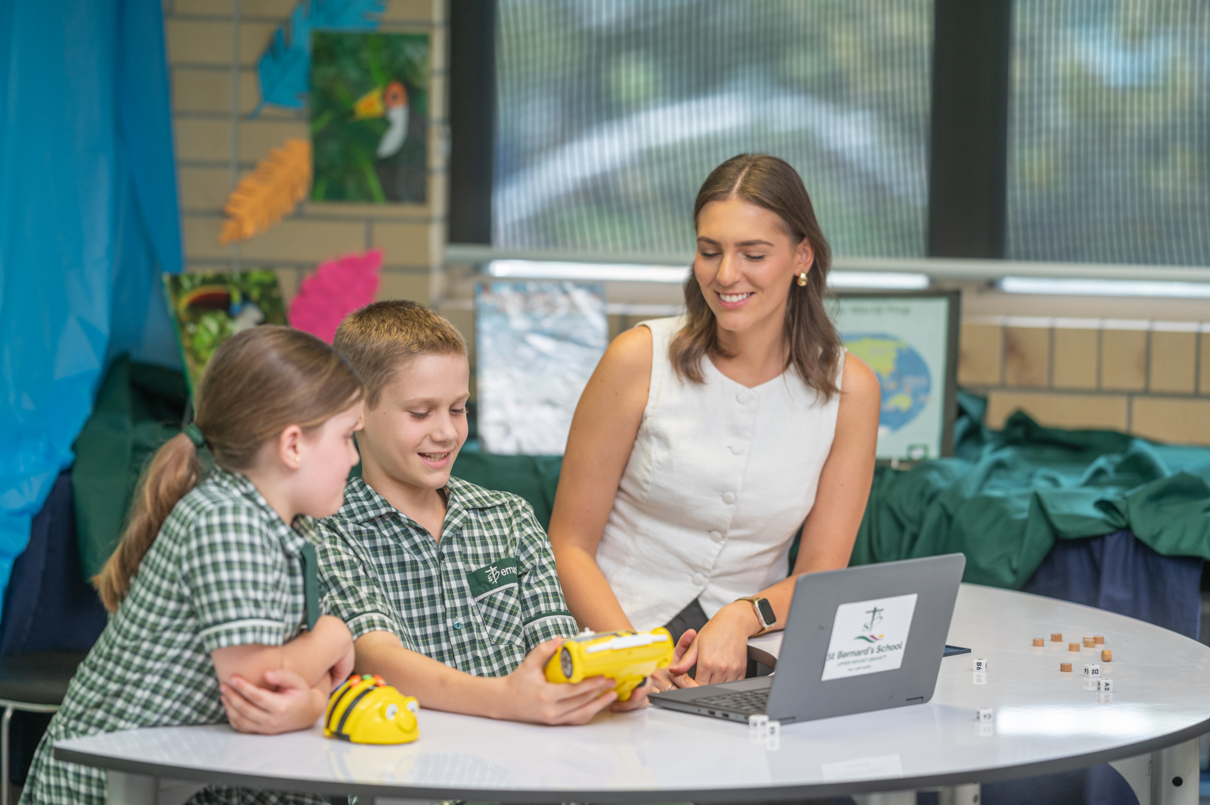 Adult supporting two students using a small device at a classroom table, with a laptop beside them.