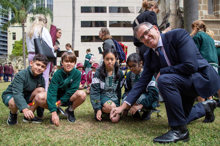 Small group of students crouching on grass with an adult in a suit, looking at something on the ground during an outdoor activity.