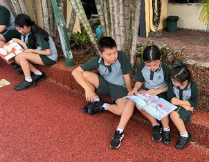 Students sitting outdoors on a bench and ground reading a book together near trees and school buildings.