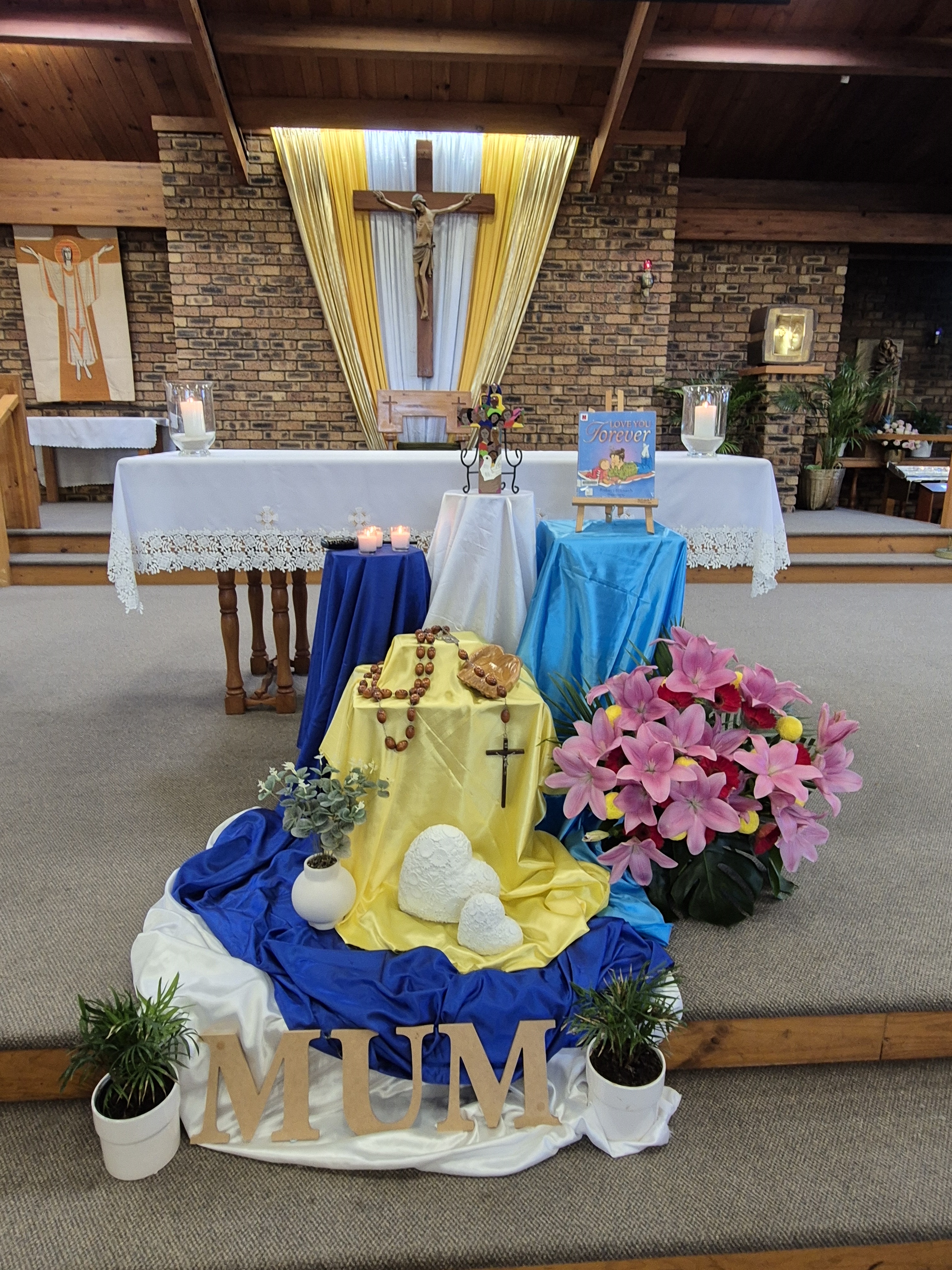 Church altar display with a central crucifix, candles, rosary beads, flowers, and draped coloured cloths, with “MUM” letters arranged at the front.