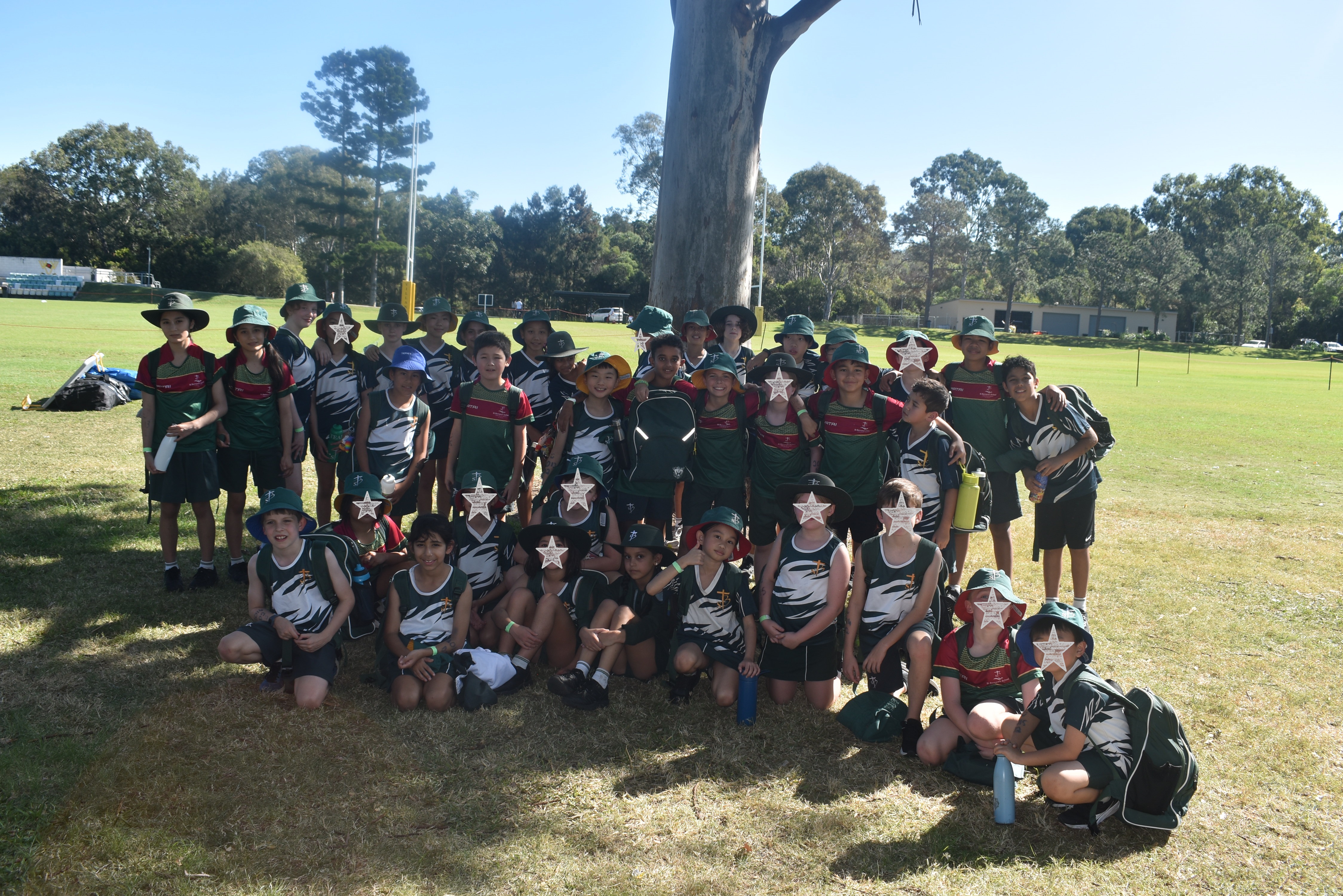 Large group of students in sports uniforms gathered on a grassy field for a group photo under a tree.